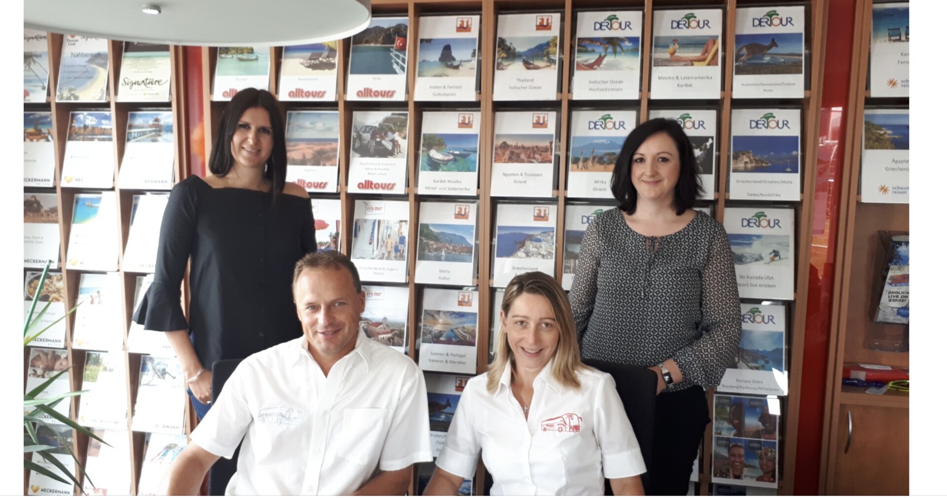 Das Team von Donaumoos Reisen vor der Katalogwand mit alltours- und DERTOUR-Reisekatalogen — zwei Reiseberaterinnen stehend, Helmut Seitz und eine Reiseberaterin in weißen Hemden sitzend